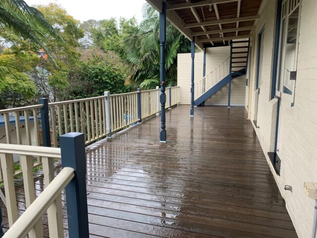 Covered timber deck with wet boards, balustrade and external stairs beside leafy garden.