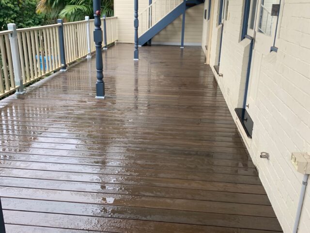 Wet timber verandah with light balustrade, blue posts, and external staircase beside white brick wall.