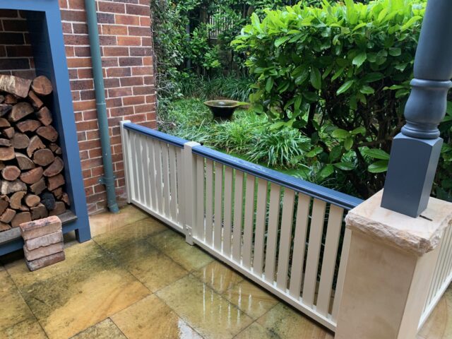 Painted timber verandah balustrade with blue handrail, wet paving, brick wall and garden.