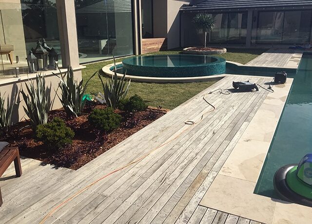 Timber pool deck with circular spa, travertine paving, landscaping, and tools indicating work in progress.