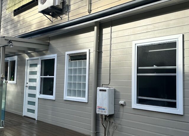Clad exterior with deck, white windows, glass-panel door, and wall-mounted hot water unit.