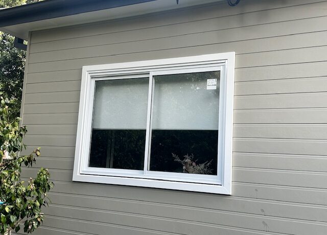 White-framed sliding window in light grey weatherboard cladding, gutter above and garden shrub at left.