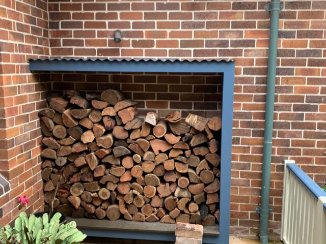 Roofed firewood storage alcove with steel frame against brick wall and paver patio.