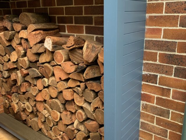 Outdoor covered firewood rack with corrugated roof against a brick wall.
