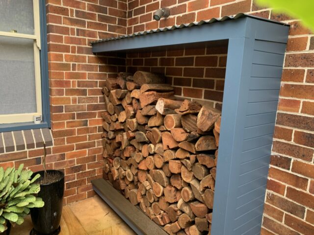 Covered log store with corrugated roof, stacked firewood against brick wall patio.